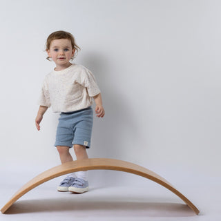 Toddler in blue shorts and cream t-shirt, standing beside a wooden wobble board