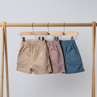 Children's twill shorts with pockets and rolled hem hanging on a rail in oat, dusty pink and slate blue