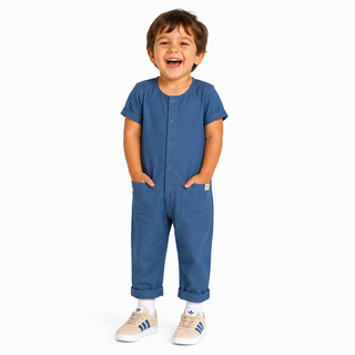 A young child wearing a short-sleeve blue overall, with front pockets, standing against a plain white background.