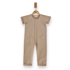 A short-sleeve one-piece romper in a an ecru colour, hanging on a wooden hanger against a plain white background. The overall features a snap-button front, two large front patch pockets, and a small white brand tag on the pocket.