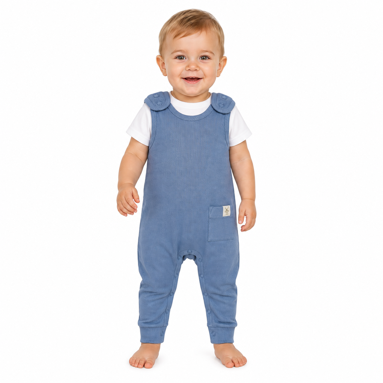 A smiling baby standing barefoot on a white background, wearing blue dungarees over a white short-sleeve shirt.