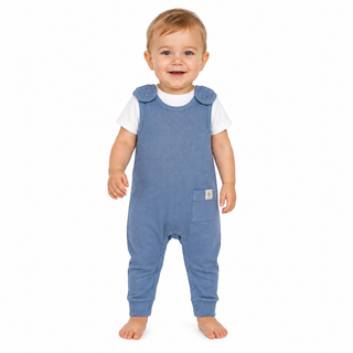 A smiling baby standing barefoot on a white background, wearing blue dungarees over a white short-sleeve shirt.