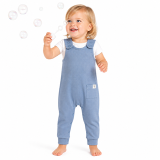 A smiling toddler stands barefoot on a white background, holding a bubble wand with floating bubbles around them. The child is wearing a light blue dungaree romper over a white short-sleeve shirt.
