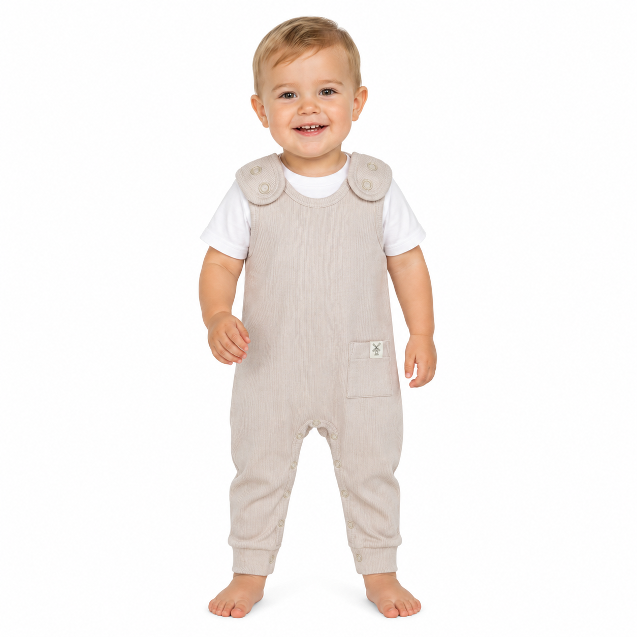 A smiling toddler standing barefoot against a plain white background, wearing light beige sleeveless dungaree over a white short-sleeve shirt.