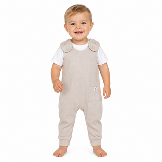 A smiling toddler standing barefoot against a plain white background, wearing light beige sleeveless dungaree over a white short-sleeve shirt.