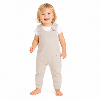 A smiling toddler standing barefoot against a white background, wearing sleeveless ecru dungarees over a white short-sleeve top.