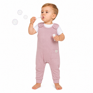 A toddler standing barefoot on a white background, wearing dusty pink overall-style baby jumpsuit over a white short-sleeve shirt. The child is blowing bubbles with one hand while holding a small bubble bottle in the other.