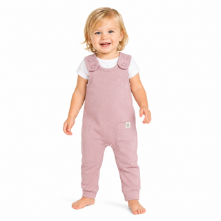 A smiling toddler standing barefoot against a plain white background, wearing dusty pink overall-style dungarees over a white short-sleeve top.