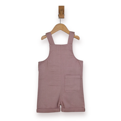 A dusty rose pink cotton short dungarees for toddlers, hanging on a wooden hanger from a wall hook against a white background.