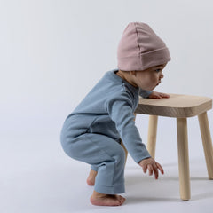 Baby dressed in a blue ribbed cotton jumpsuit and pink beanie hat, crouching down while using a wooden stall for balance.