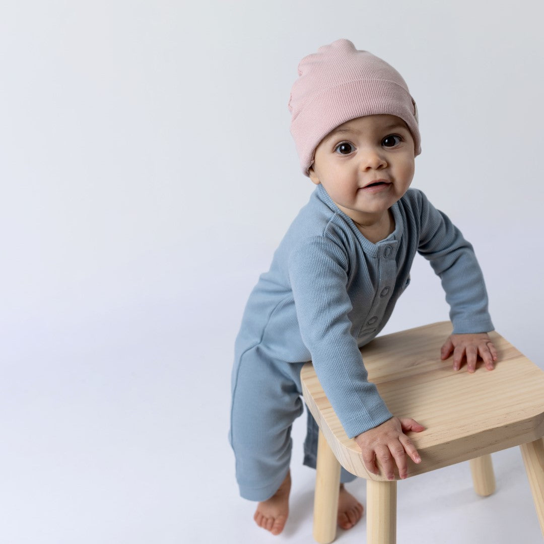 Baby wearing a blue ribbed cotton jumpsuit and pink beanie hat, standing upright while holding onto a wooden stall for support, looking directly at the camera.