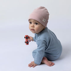 Baby girl wearing a blue ribbed cotton jumpsuit and a pink beanie hat, sitting on a neutral-toned floor while playing with a wooden toy car.