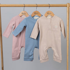 A set of three ribbed cotton baby jumpsuits in neutral tones; blue, oat and pink, hanging on wooden hangers from a wooden clothing rail.