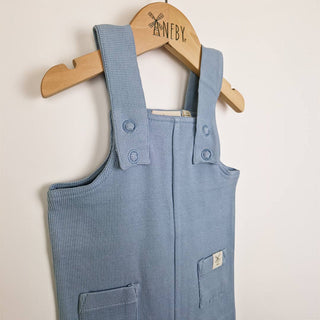 Close up view of a pair of baby ribbed cotton dungarees in slate blue on a wooden hanger showing adjustable shoulder straps and neat stitching around the armhole and neckline.