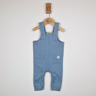 Front view of a pair of baby ribbed cotton dungarees in slate blue on a wooden Aneby coat hanger showing adjustable shoulder straps with blue metal poppers and square patch pockets.  