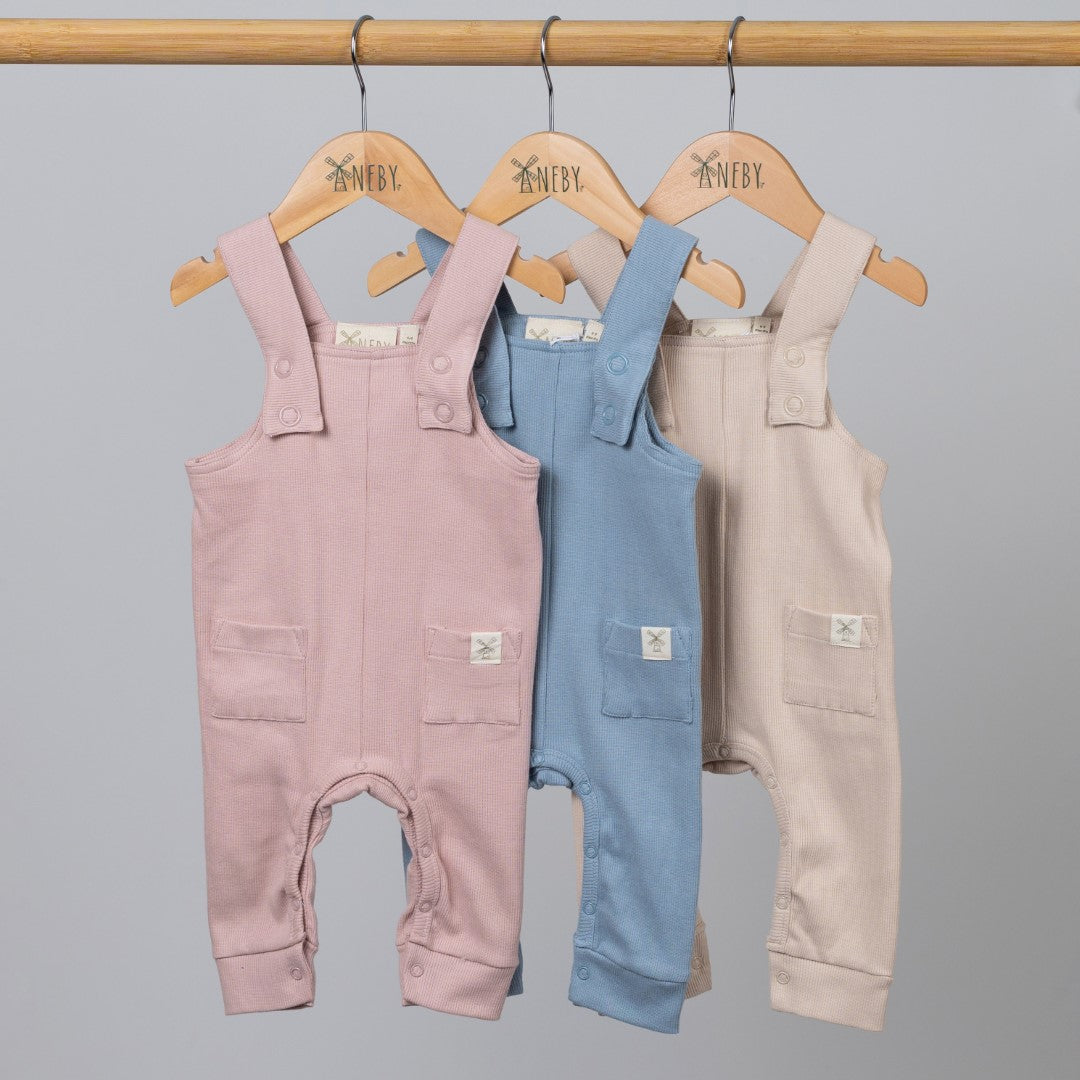 Three pairs of baby ribbed cotton dungarees in slate blue, dusty pink, and oat, neatly arranged on a wooden rail with wooden Aneby coat hangers. The image highlights the distinct colours and detailed ribbed fabric.