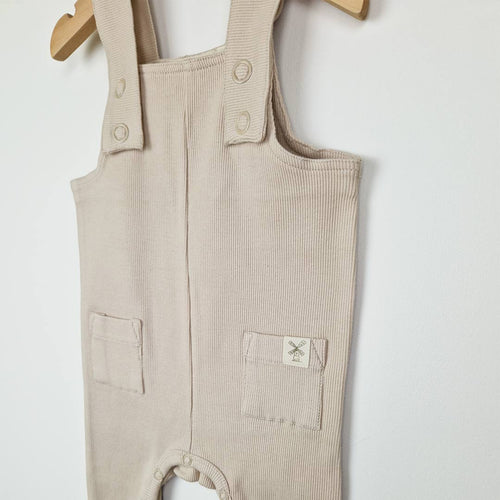 Close up view of a pair of baby ribbed cotton dungarees in oat on a wooden hanger showing ribbed shoulder strap with oat colour metal ring poppers. 
