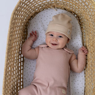 Small baby smiling while lying in a moses basket, dressed in a dusty pink sleeveless rib baby romper and a oat beanie hat.