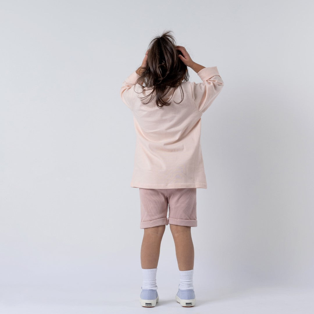 Girl with her back to the camera wearing a girls dusty pink long sleeve t-shirt and pink shorts. Her hands rest on top of her head as she stands against a plain background, highlighting the t-shirt's back design and fit.