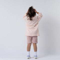 Girl with her back to the camera wearing a girls dusty pink long sleeve t-shirt and pink shorts. Her hands rest on top of her head as she stands against a plain background, highlighting the t-shirt's back design and fit.