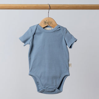 Slate blue short sleeve baby bodysuit hanging neatly on a wooden hanger, made from ribbed organic cotton with soft rounded shoulders.