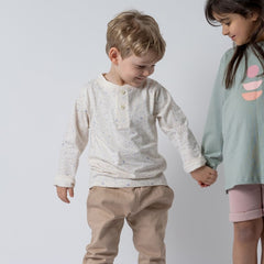 Smiling young boy in a kids long sleeve ecru t-shirt and oat chinos holding hands with a girl, sharing a joyful moment against a plain background.