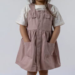 A close-up of a young girl wearing an ecru children's paint splat t-shirt underneath a dusty pink pinafore dress. The colourful speckled design is visible on the t-shirt’s sleeves and neckline, subtly peeking out from under the dress.