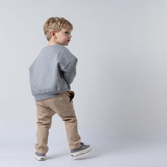 Small blonde boy with his back to the camera, wearing a grey children's spaceman sweatshirt and oat chinos with a back pocket hem tag. Looking off into the distance. Neutral background.