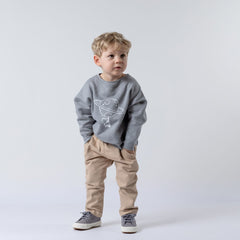 Small blonde boy wearing a grey children's spaceman sweatshirt, hands in pockets, looking off into the distance. The sweatshirt features an astronaut print with a planet and stars. Neutral background.