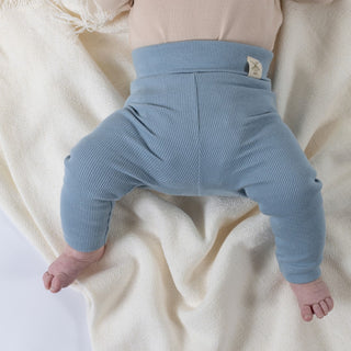 Close-up of organic ribbed slate blue baby leggings resting on a cream blanket, highlighting the fold-over waistband with the Aneby windmill hem tag visible. The fine ribbed texture and high-quality finish are evident.
