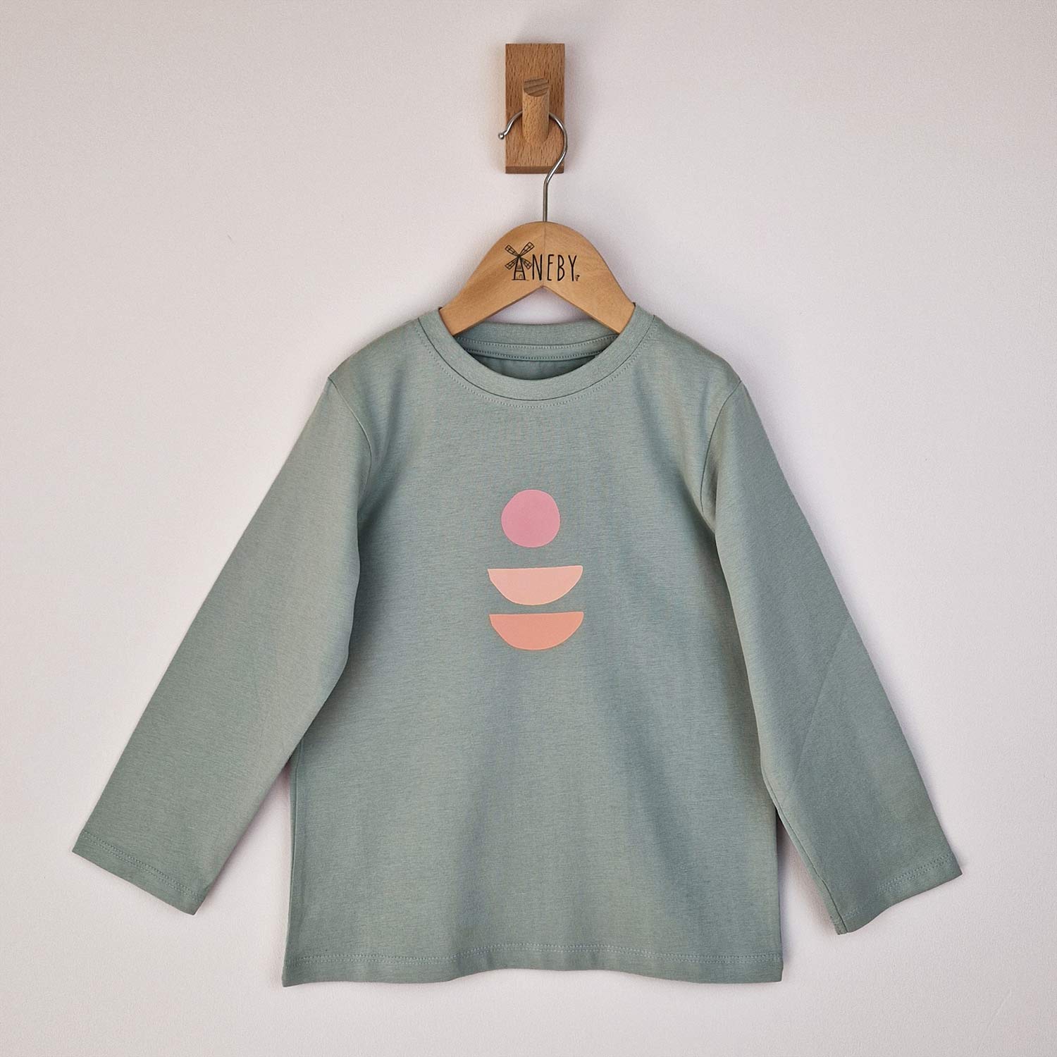 lat lay of a children's sage green long sleeve t-shirt displayed on a neutral background. The shirt features a simple round neckline, relaxed fit and abstract logo on the front. 