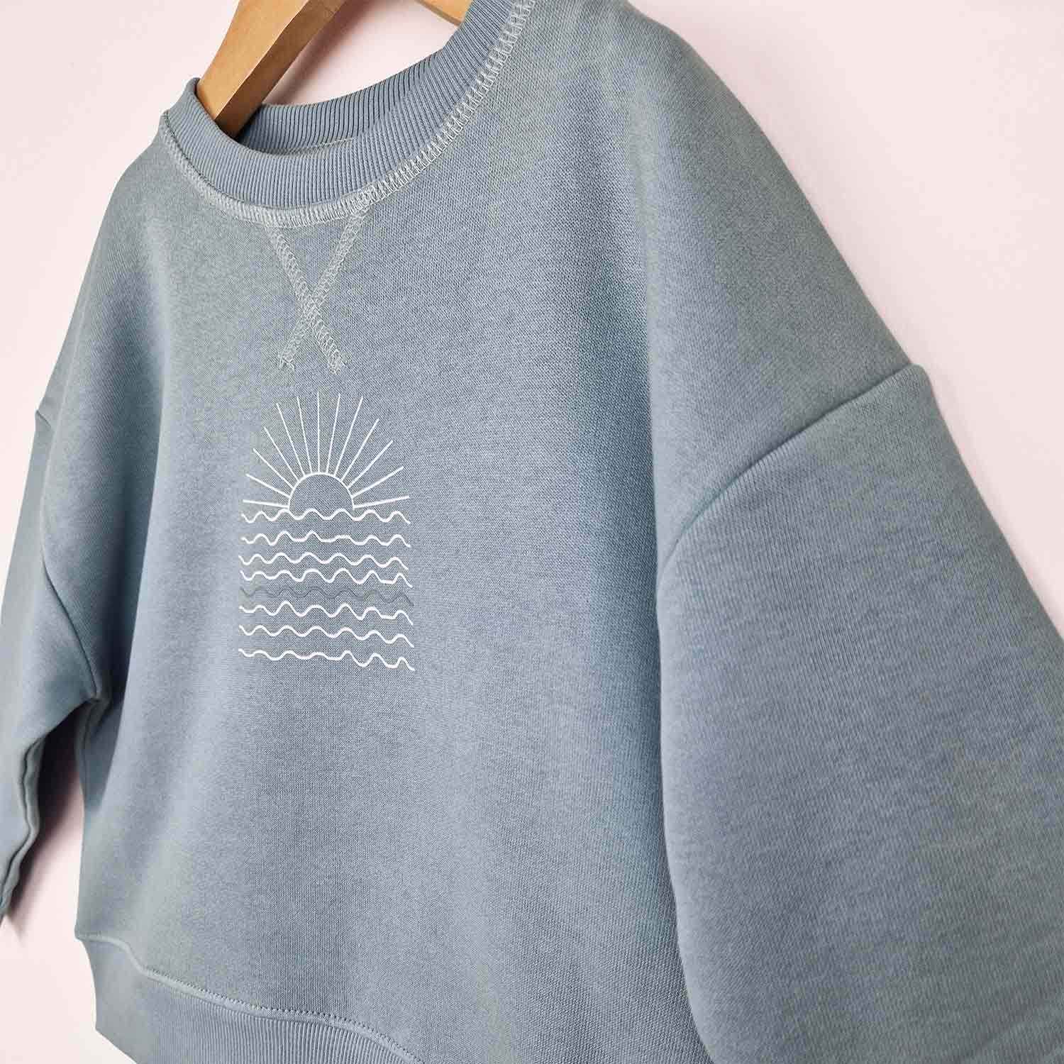 Flat lay of a steel blue sweatshirt featuring a white sunrise graphic on the front, displayed against a neutral background.