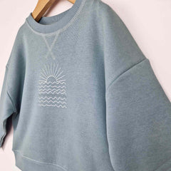 Flat lay of a steel blue sweatshirt featuring a white sunrise graphic on the front, displayed against a neutral background.