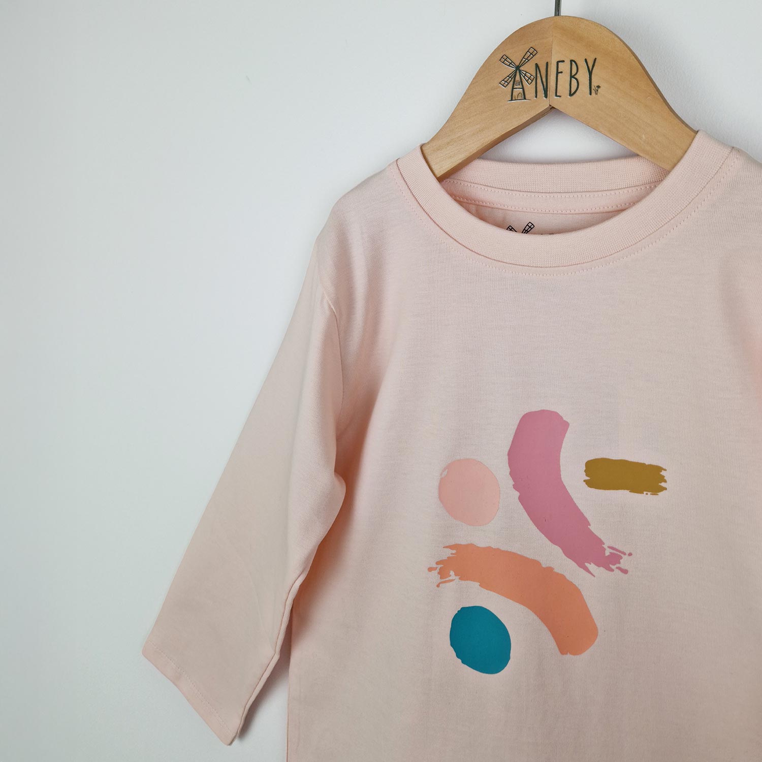 Close up of a girls dusty pink long sleeve t-shirt showing the front jersey neckline with neat edge stitching and the bold paint dash print on the front in shades of pink, mustard and turquoise.   