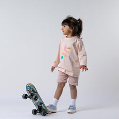 Girl standing on a skateboard wearing a girls dusty pink long sleeve t-shirt and matching pink Aneby shorts. She is looking to the side against a neutral background
