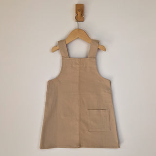 Back of an oat-coloured organic cotton dungaree dress on a wooden hanger, showing the shoulder straps, single patch pocket on right side and minimalistic design.