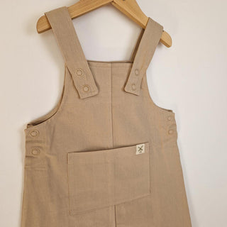 Close up front of an oat-coloured organic cotton dungaree dress, highlighting the front pocket, sturdy straps with oat coloured metal rng snaps and high quality soft twill fabric.