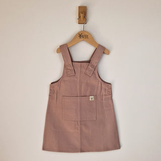 A dusty pink organic cotton dungaree dress on a wooden hanger, showing the front details including the large patch pocket with Aneby windmill hemtag and poppered straps.