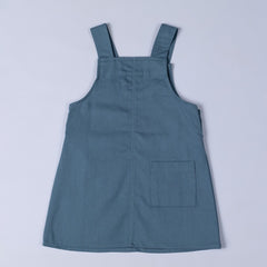 Flat lay of the back of a slate blue organic cotton dungaree dress, with back straps and a simple, timeless design.