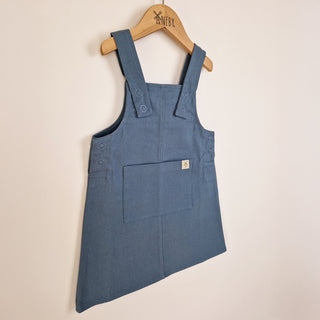 A slate blue organic cotton dungaree dress, featuring a large front kangaroo pocket and classic dungaree-style straps.
