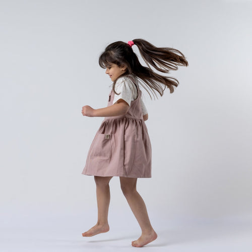 A dusty pink girls’ pleated dress with straps, displayed on a child. The dress features a flowing pleated skirt and delicate straps, perfect for special occasions.