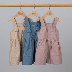 A set of three girls’ pleated dresses with straps in slate blue, dusty pink, and oat, arranged together to showcase the available colours.