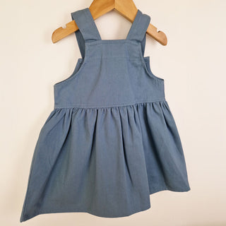 Flat lay of the back of a slate blue girls’ pleated dress with straps, focusing on the pleats and back strap details.