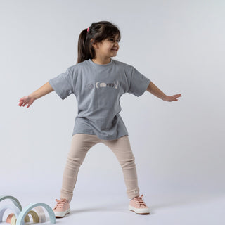 A young girl wearing a kids' abstract t-shirt with oat-coloured leggings, standing with her arms stretched wide. The t-shirt features a geometric pattern in earthy tones.