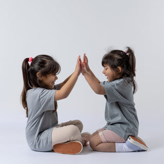 Two young girls sitting on the floor, side-on, wearing kids' abstract t-shirts. They are laughing and double high-fiving each other.