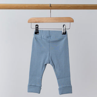 A back view of slate blue ribbed leggings hanging from a wooden frame. The fabric texture is clearly visible, with the elasticated waistband slightly gathered. The background remains neutral.