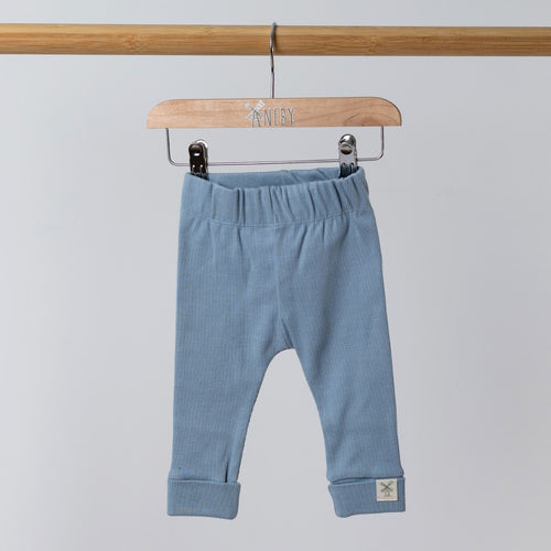A pair of slate blue ribbed leggings hangs from a wooden frame. The Aneby hem tag is visible on the cuff. The elasticated waist is slightly gathered, and the background is a neutral tone.