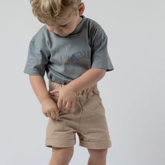 A young boy with shaggy blonde hair wearing a blue-grey kids' camping t-shirt tucked into oat-coloured chino shorts. He is placing an item into his pocket.