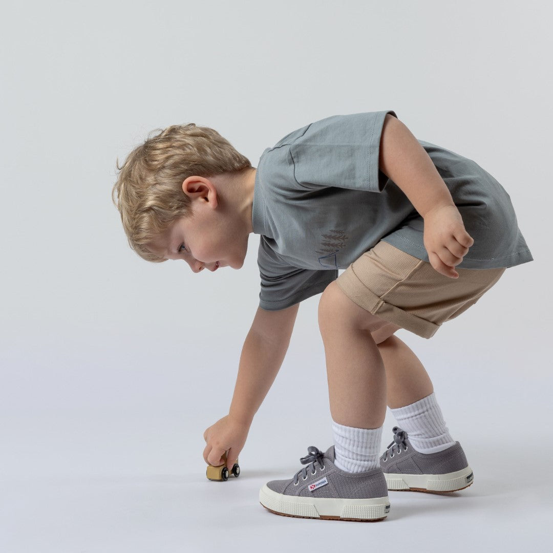 A young boy in a blue-grey kids' camping t-shirt and oat-coloured chino shorts, leaning over while playing with a small wooden toy car.