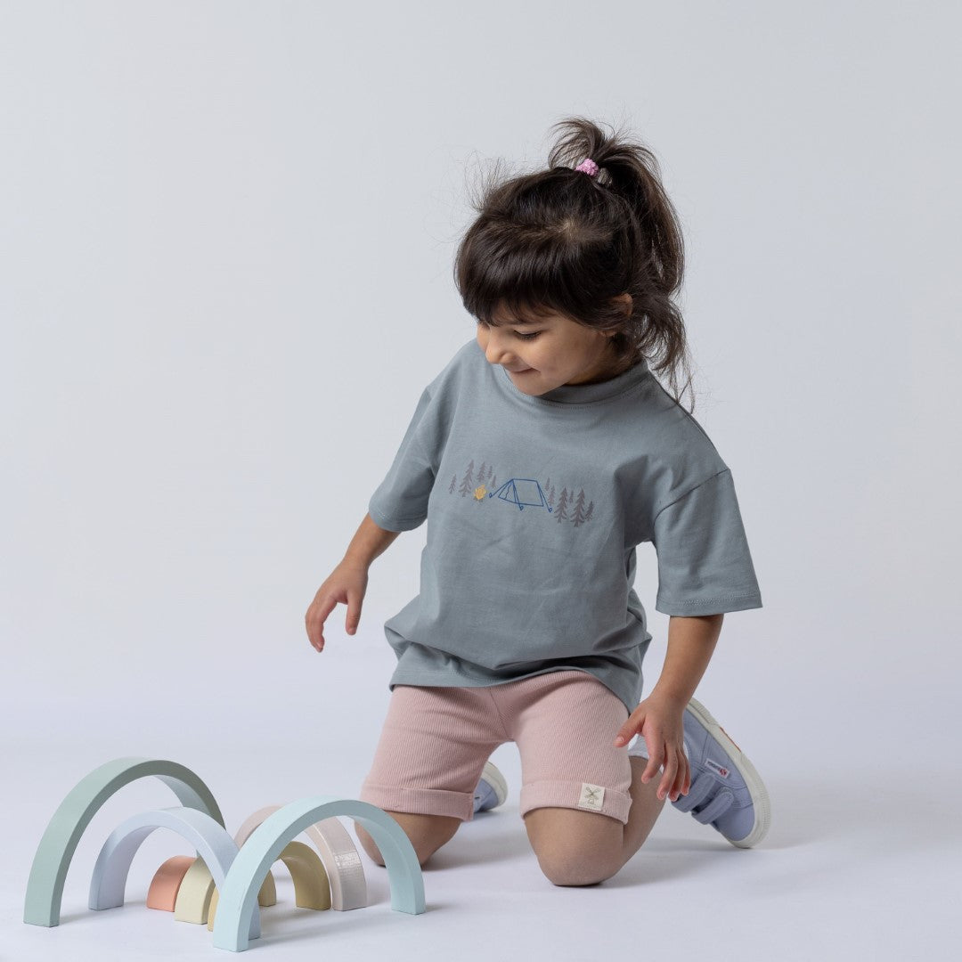 A young girl wearing a kids' camping t-shirt with pink shorts, playing with classic wooden toy shapes on the floor. The t-shirt features a tent, campfire, and fir trees design.